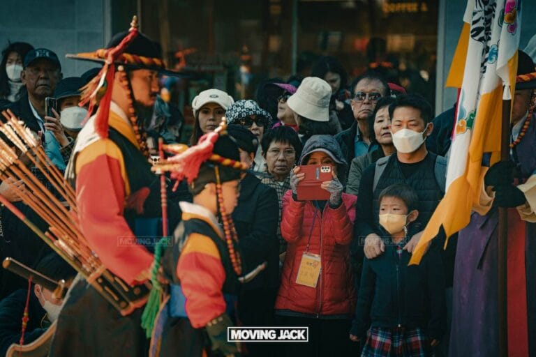A crowd enjoys a traditional cultural parade—one of the free things to do in seoul—with two people in colorful historical costumes. Some spectators wear masks, while another records the vibrant scene with a red phone. -copyright-moving-jack. Com