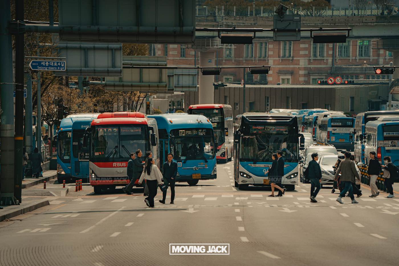 Bezoek Seoul: 12 fascinerende eerste indrukken die je meteen zult herkennen 11 Seoul Drukke stadsstraat met meerdere blauwe en rode bussen die stoppen terwijl voetgangers de kruising oversteken; stedelijke gebouwen en viaducten op de achtergrond, die het dagelijks leven en enkele gratis dingen om te doen in Seoul benadrukken. -copyright-moving-jack.com