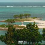 A small, sandy island with a few clusters of trees, a lifeguard tower, and clear turquoise water, viewed from a higher vantage point with greenery in the foreground. -copyright-moving-jack. Com