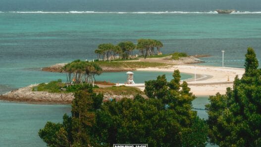 A small, sandy island with a few clusters of trees, a lifeguard tower, and clear turquoise water, viewed from a higher vantage point with greenery in the foreground. -copyright-moving-jack. Com