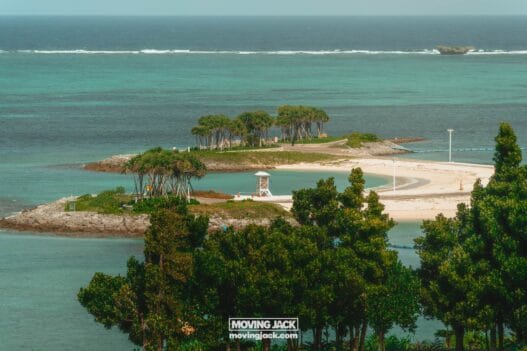 Een klein, zanderig eiland met een paar groepjes bomen, een reddingspost en helder turquoise water, gezien vanaf een hoger uitkijkpunt met groen op de voorgrond. -copyright-moving-jack.com