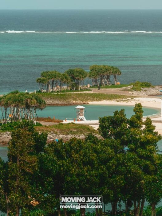 Een klein, zanderig eiland met een paar groepjes bomen, een reddingspost en helder turquoise water, gezien vanaf een hoger uitkijkpunt met groen op de voorgrond. -copyright-moving-jack.com