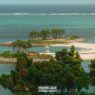 A small, sandy island with a few clusters of trees, a lifeguard tower, and clear turquoise water, viewed from a higher vantage point with greenery in the foreground. -copyright-moving-jack. Com