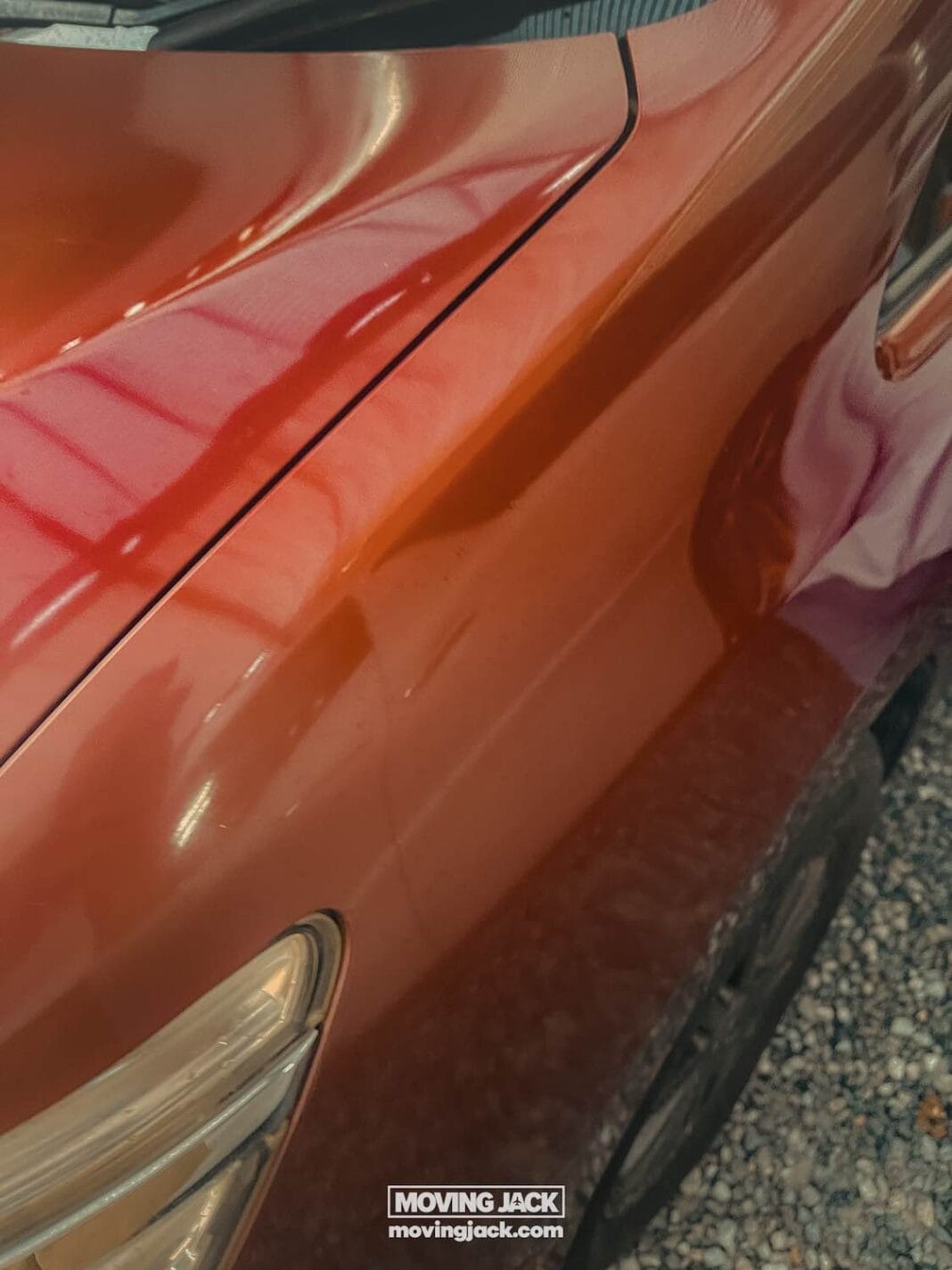 Close-up of an orange car’s front side, showing the fender, headlight, and part of the hood—perfect inspiration for renting a car in okinawa. The polished surface reflects a nearby structure as it rests on gravel. -copyright-moving-jack. Com