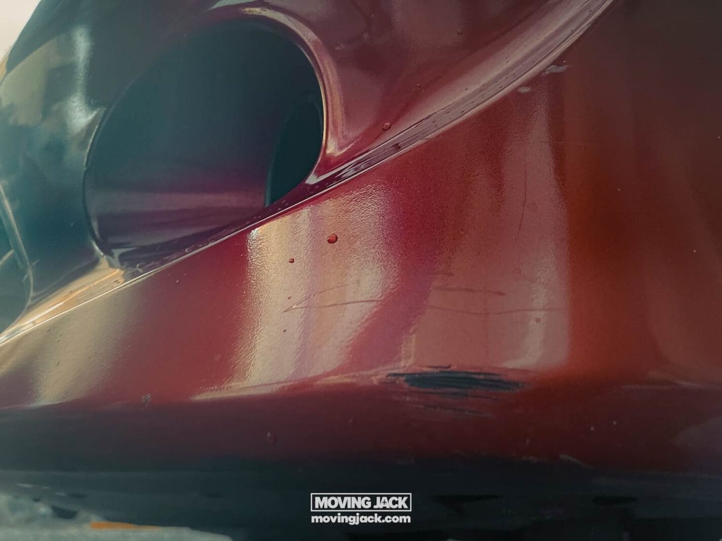 Close-up of a red car bumper with visible scratches and scuff marks, possibly from renting a car in okinawa. The bumper has a round hole, a reflective surface, and a "moving jack" sticker at the bottom. -copyright-moving-jack. Com