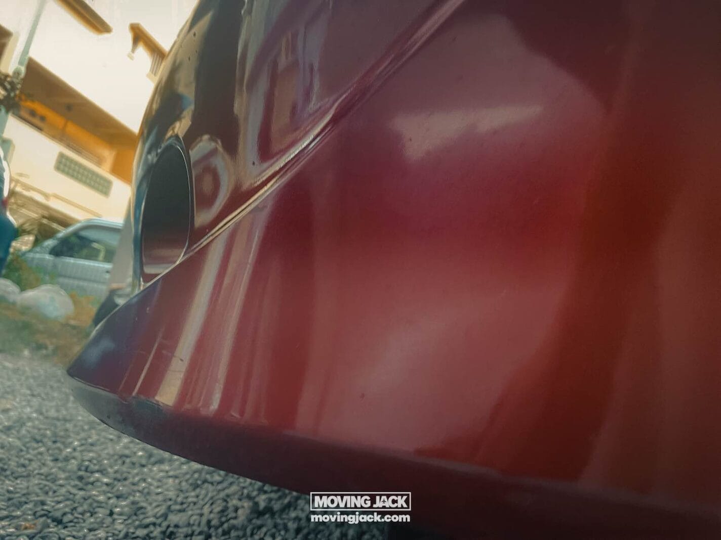 Close-up view of the rear bumper of a red car parked on gravel, with part of another car and buildings visible. A "moving jack" sticker is on the bumper—a familiar sight when renting a car in okinawa. -copyright-moving-jack. Com