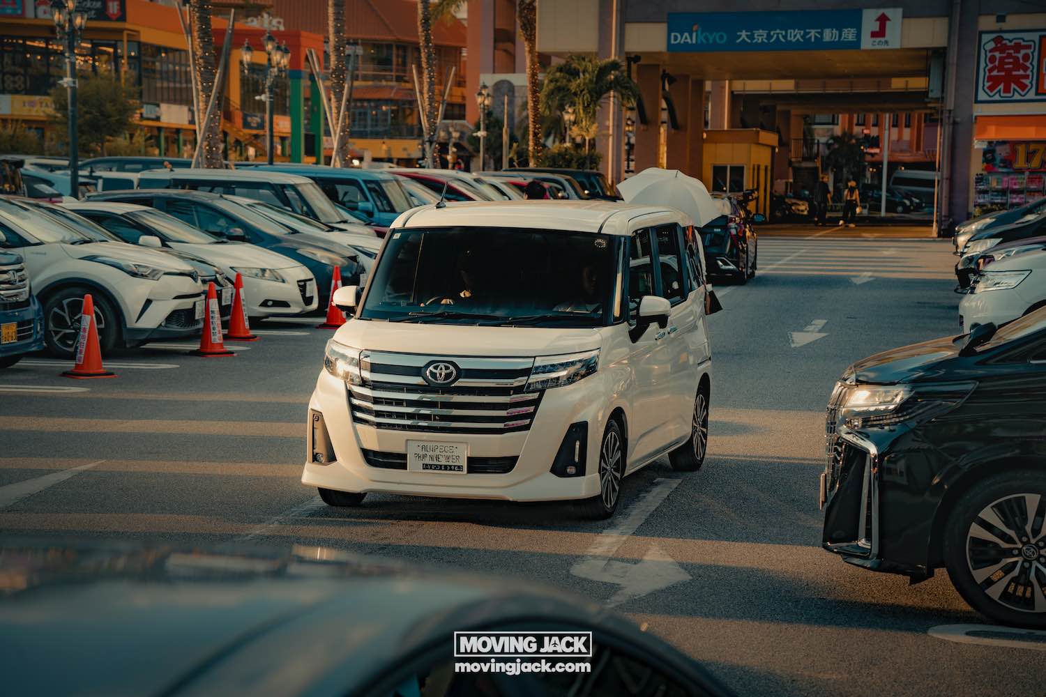A white toyota van, perfect for renting a car in okinawa, drives through a busy parking lot surrounded by parked cars, with shops and japanese signage visible in the background. -copyright-moving-jack. Com