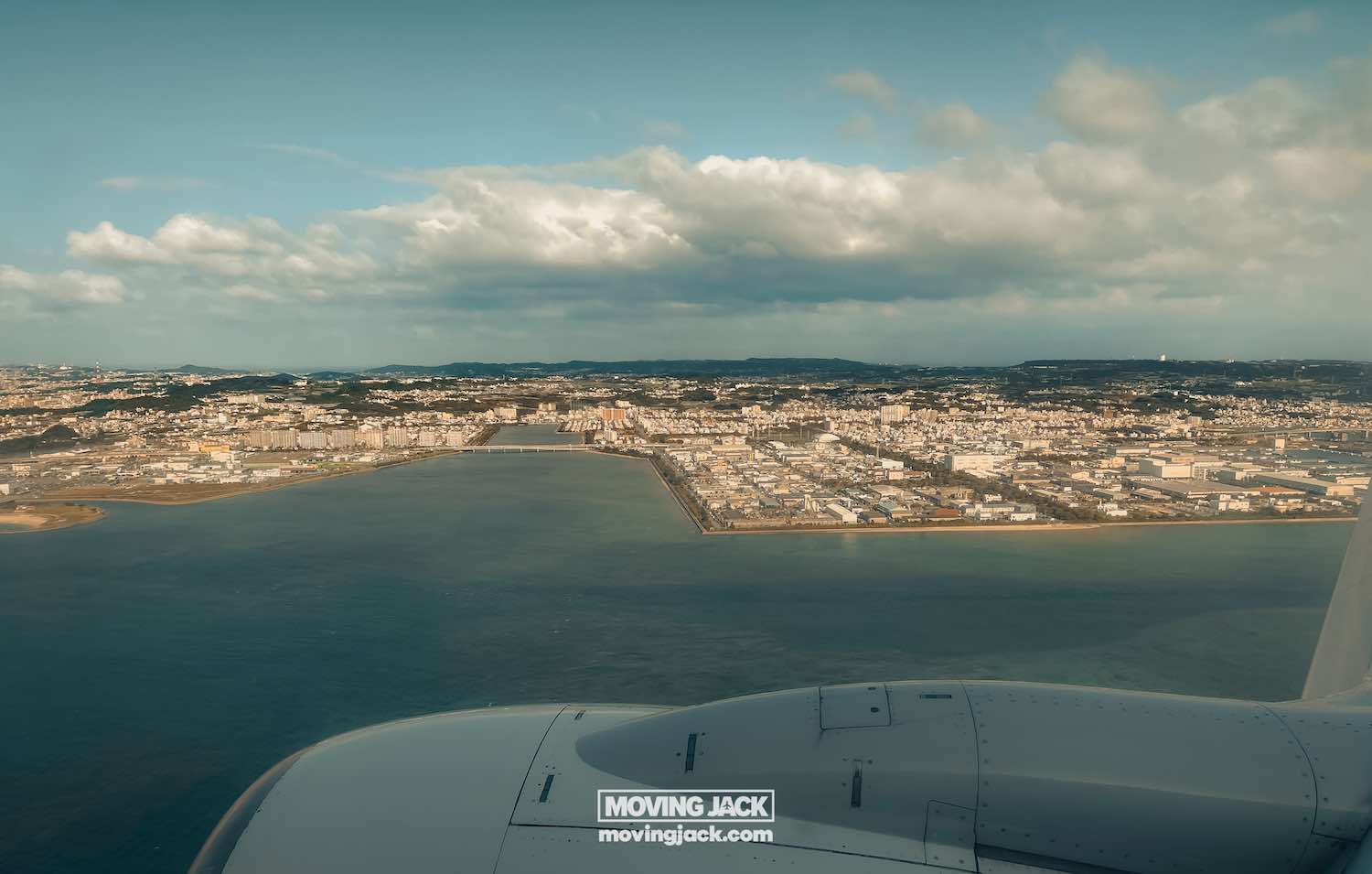 Aerial view from an airplane window showing a city bordered by water with a partly cloudy sky—perfect for exploring after landing and renting a car in okinawa. Part of the plane's wing and engine are visible in the foreground. -copyright-moving-jack. Com