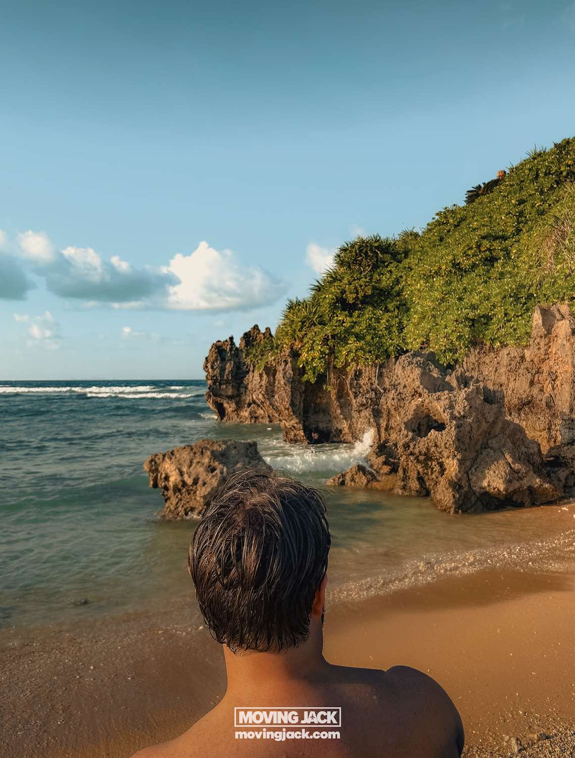 A person with short brown hair stands on a sandy beach in okinawa, facing rocky cliffs covered in green vegetation—waves crash ashore beneath a blue sky. Renting a car in okinawa makes exploring such scenic coastal views effortless. -copyright-moving-jack. Com