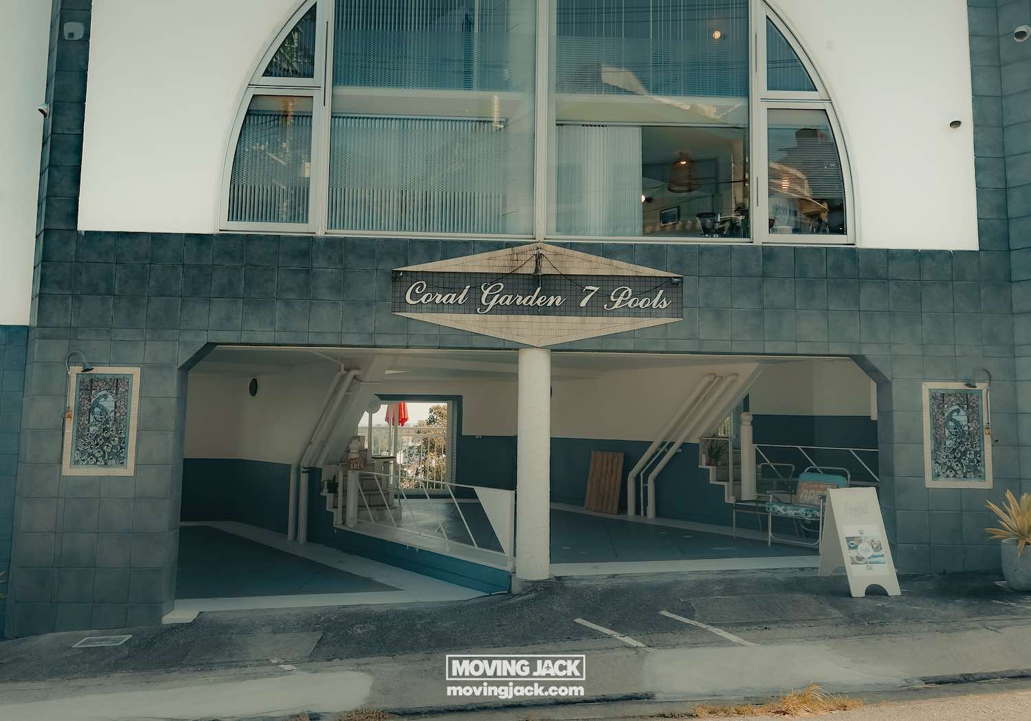Entrance to a modern building with a sign reading "coral garden 7 pool," featuring large windows, a covered driveway, and a sandwich board sign. Perfect spot for visitors renting a car in okinawa, with gray exterior walls and white accents. -copyright-moving-jack. Com