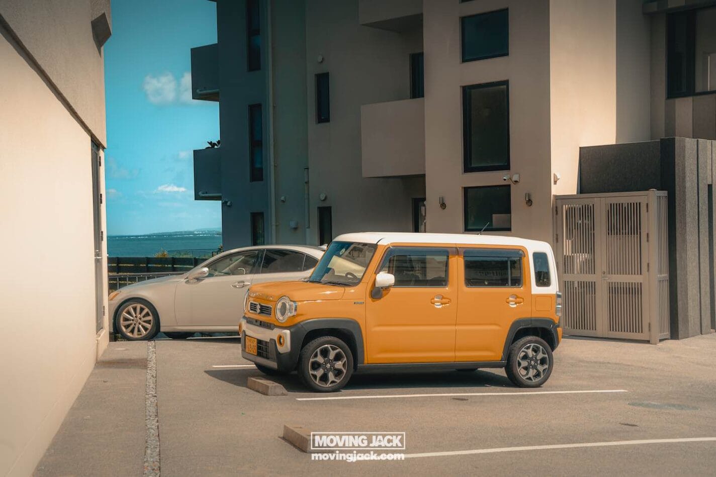 A yellow and white compact suv is parked next to a silver sedan in a modern apartment parking lot on a sunny day, with blue sky and ocean visible in the background. -copyright-moving-jack. Com