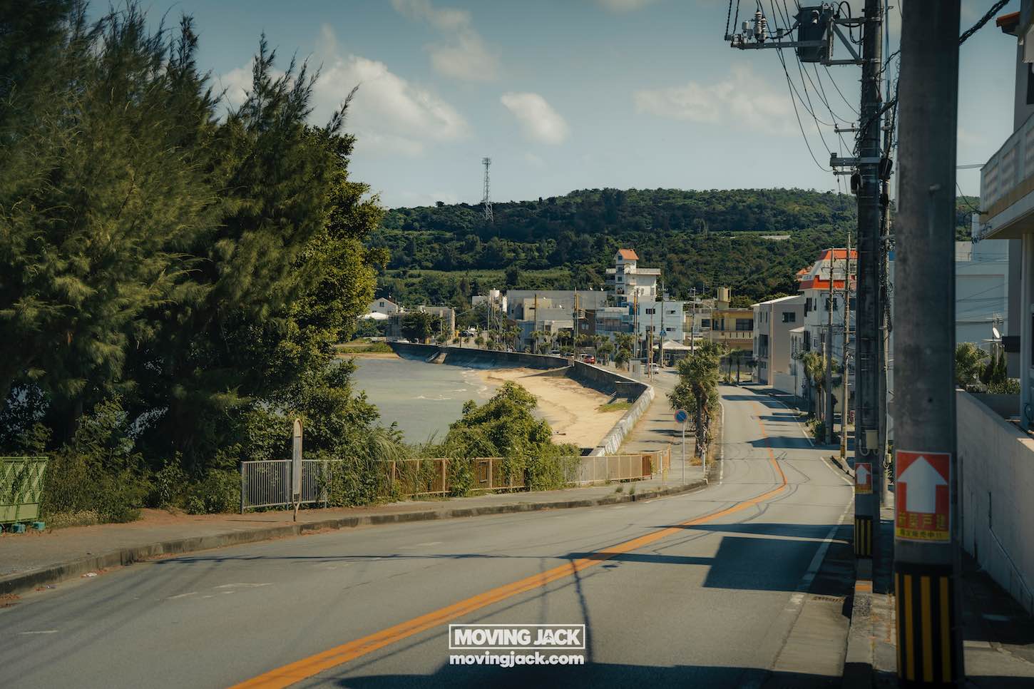 A coastal road, perfect for renting a car in okinawa, curves alongside a sandy beach and blue water, bordered by trees, utility poles, and buildings under a partly cloudy sky. A forested hill rises in the background. -copyright-moving-jack. Com
