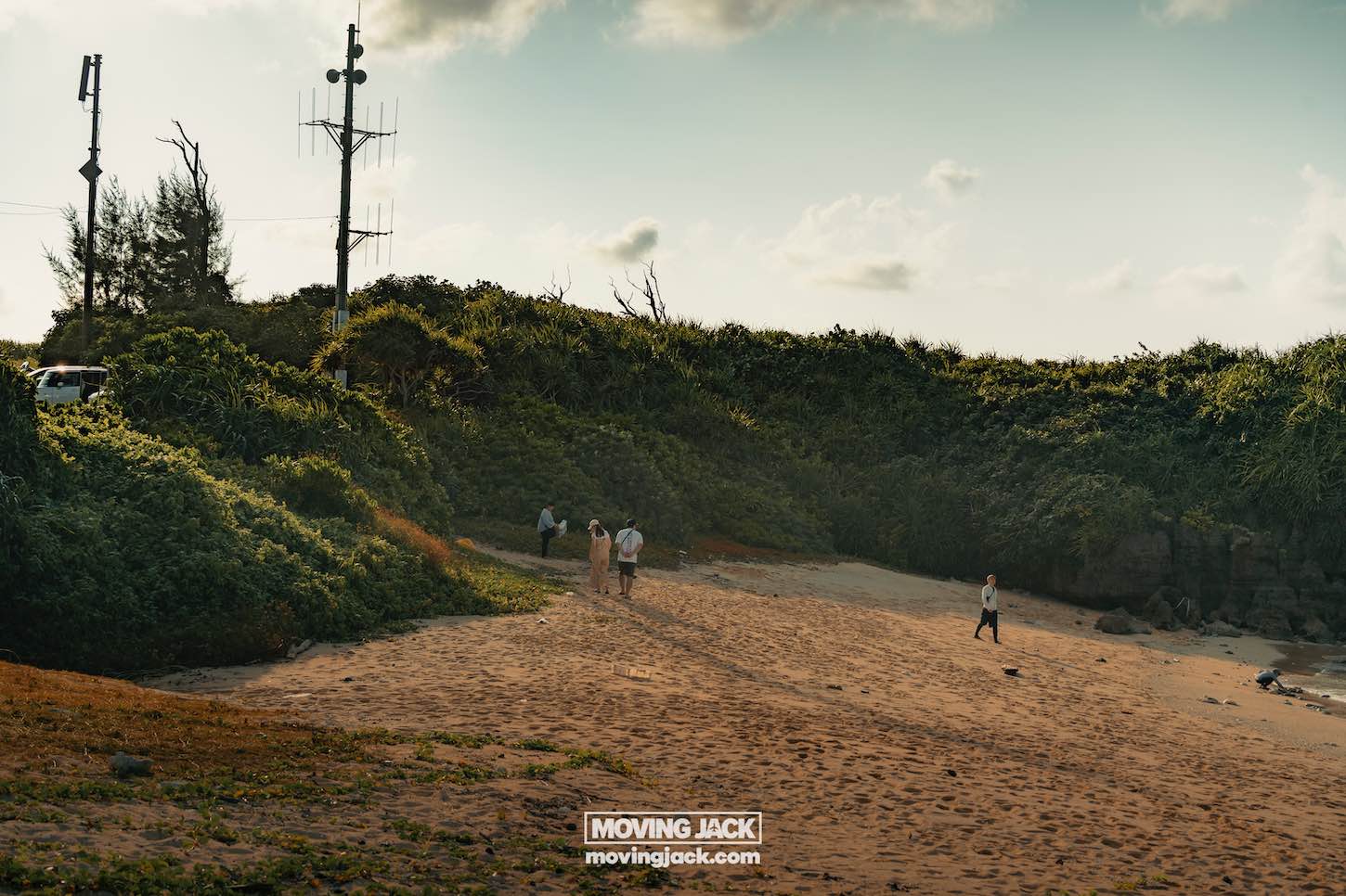 A small group walks along a sandy beach bordered by green vegetation and rocky cliffs under a partly cloudy sky. A communications tower and a parked vehicle—ideal if you’re renting a car in okinawa—are visible on the left. -copyright-moving-jack. Com