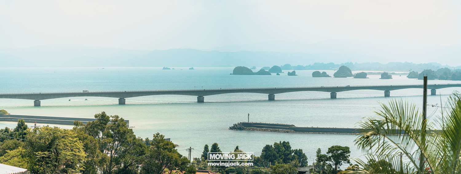 A long bridge stretches across a calm blue bay, the perfect scenery to explore when renting a car in okinawa. -copyright-moving-jack. Com