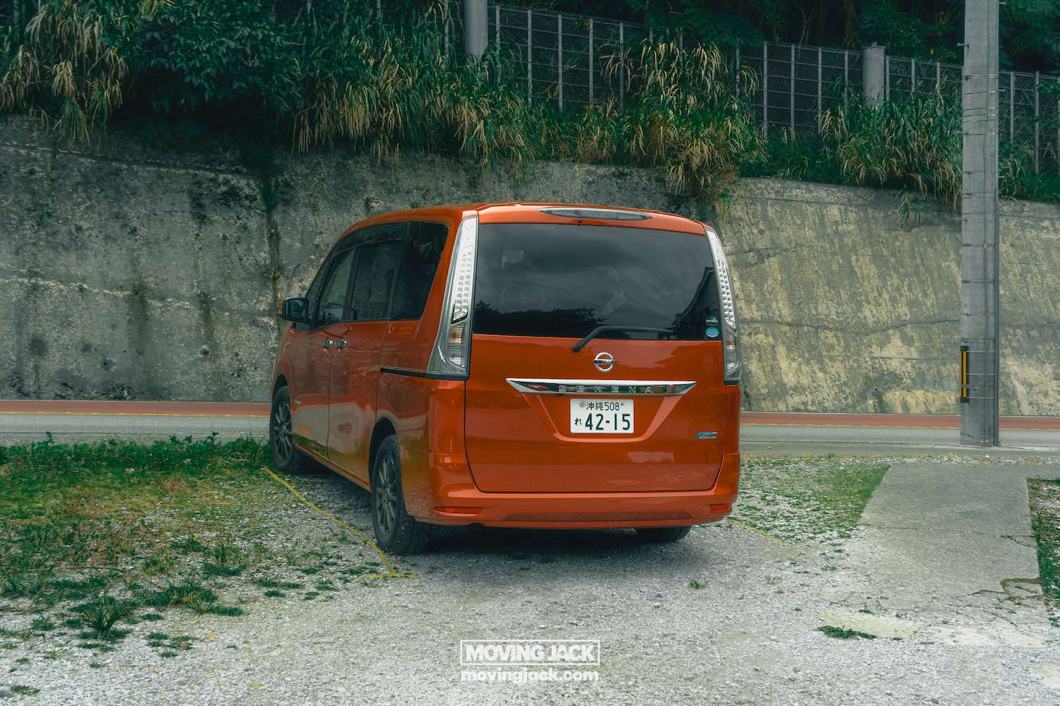 A red nissan van is parked on a gravel spot near a concrete wall with greenery above it in okinawa. Perfect for those renting a car in okinawa, the license plate reads "42-15," and a "moving jack" logo is visible on the ground behind the van. -copyright-moving-jack. Com