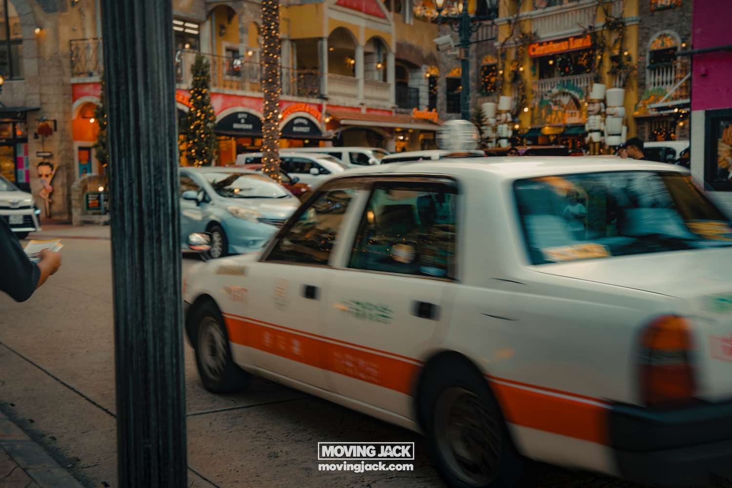 A white and orange taxi drives past shops decorated with lights and christmas trees on a busy street in okinawa—a lively scene you might spot while renting a car in okinawa. A blurred pedestrian is visible on the left and other cars are parked in the background. -copyright-moving-jack. Com