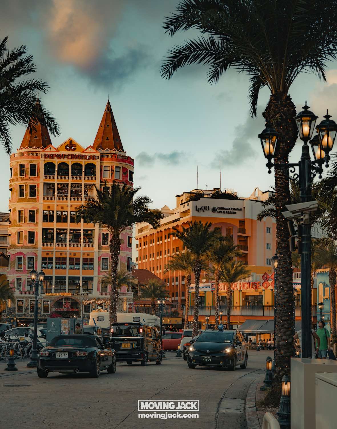 A lively street scene at sunset, with colorful, ornate buildings, palm trees, and street lamps. Cars, perhaps rented by those renting a car in okinawa, move along the road as a partly cloudy sky adds warmth to the vibrant urban atmosphere. -copyright-moving-jack. Com