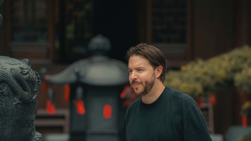 About moving jack 2 about me A man with brown hair and a beard, wearing a black shirt, stands outdoors near a decorative stone sculpture. The blurred background of a traditional building and red hanging ornaments adds depth to this casual "about me" moment. -copyright-moving-jack. Com