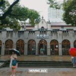 People with umbrellas stand in front of a historic building with arched windows and a "tsutaya books" sign. Trees flank the entrance and the ground glistens with rain—perfect for those seeking things to do in the french concession. -copyright-moving-jack. Com