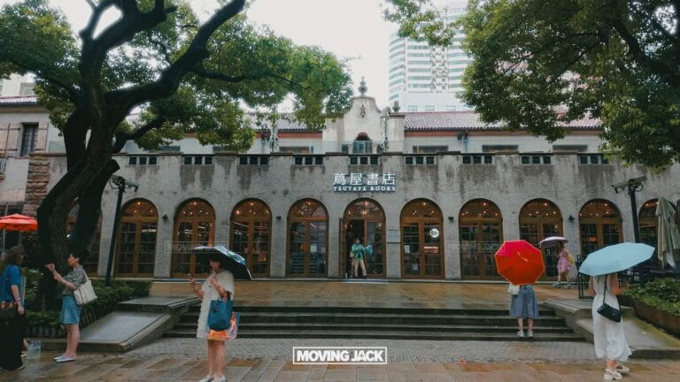 People with umbrellas stand in front of a historic building with arched windows and a "tsutaya books" sign. Trees flank the entrance and the ground glistens with rain—perfect for those seeking things to do in the french concession. -copyright-moving-jack. Com