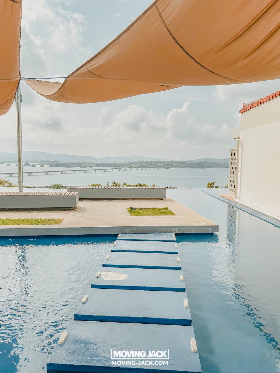 Een modern zwembad met drijvende blauwe stapstenen, beschaduwd door oranje zeildoeken, biedt uitzicht op een schilderachtig uitzicht op een baai die doet denken aan de beste stranden van Okinawa. Het merk "moving jack" is zichtbaar op de rand van het zwembad. -copyright-moving-jack.com