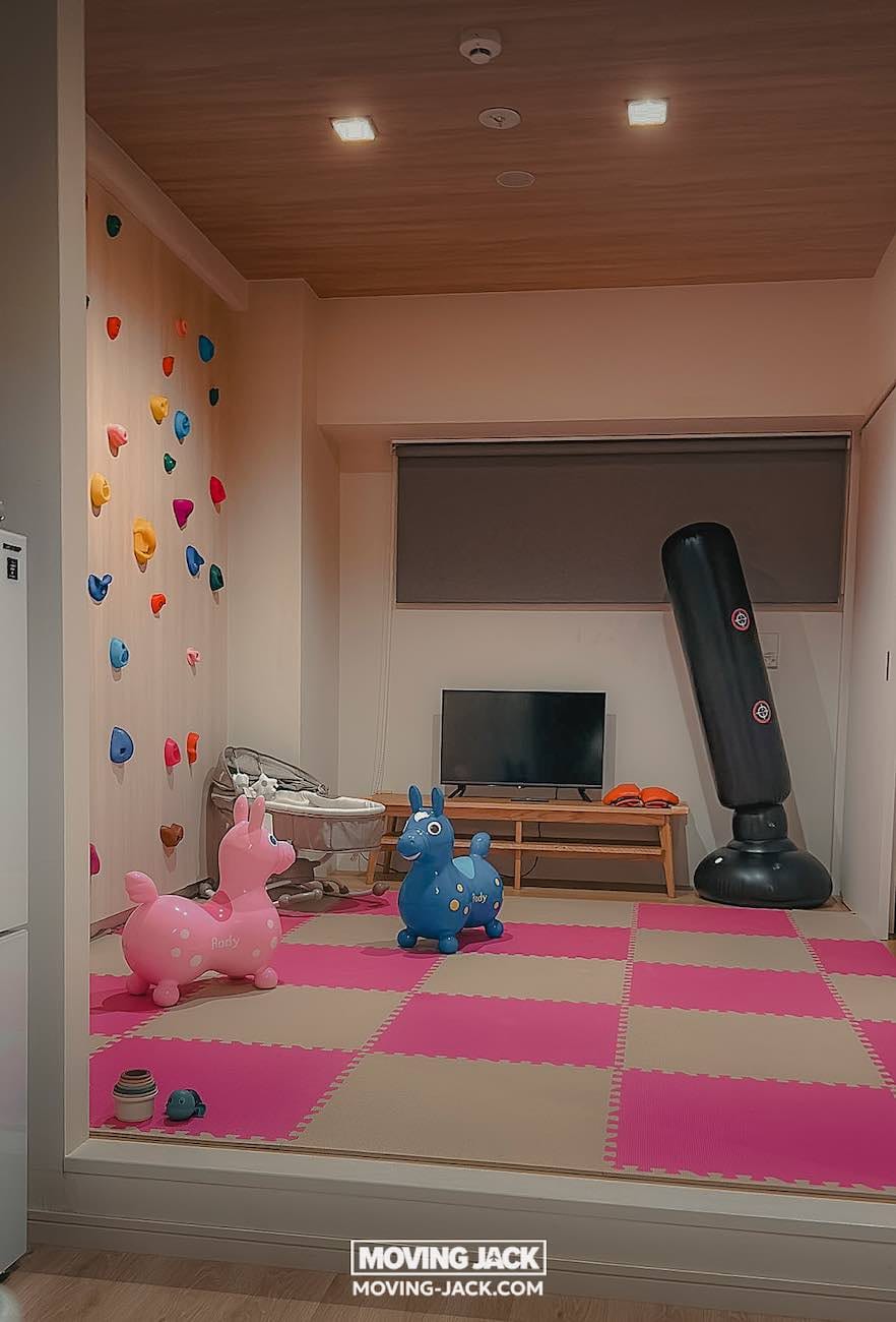 Een speelkamer voor kinderen, geïnspireerd op de levendige kleuren van de beste stranden van Okinawa, met een klimwand, een tv op een lage standaard, een boksbal, twee opblaasbare dieren en roze-beige schuimmatten. Er staat een stoeltje bij de muur. -copyright-moving-jack.com