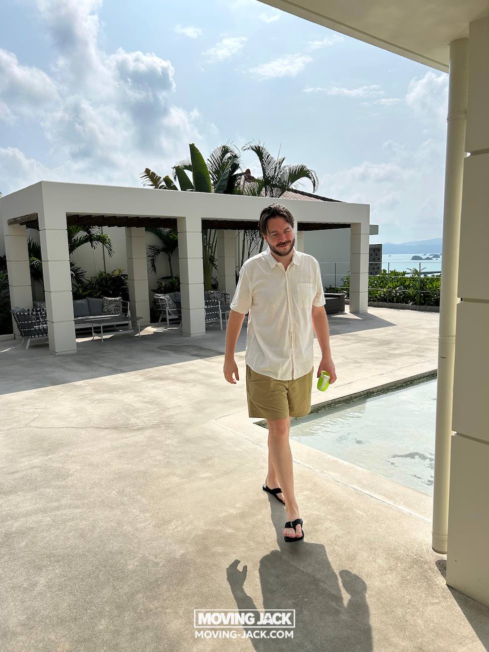 Een man in een licht overhemd en korte broek loopt langs een zwembad onder een gedeeltelijk bewolkte hemel, met een drankje in zijn hand. Terrasmeubilair en tropische planten roepen de sfeer op van de beste stranden die Okinawa te bieden heeft. Het logo "moving jack" staat onderaan. -copyright-moving-jack.com