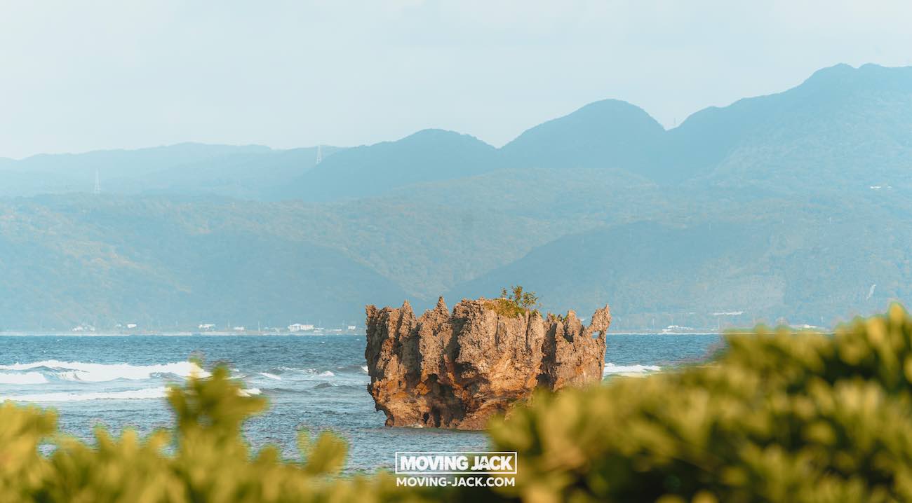 Een grillige rotsformatie rijst op uit de oceaan, bedekt met kleine planten, tegen een achtergrond van verre groene bergen en een heldere lucht – een iconisch tafereel nabij de beste stranden die Okinawa te bieden heeft, met lommerrijke struiken die de voorgrond omlijsten. -copyright-moving-jack.com