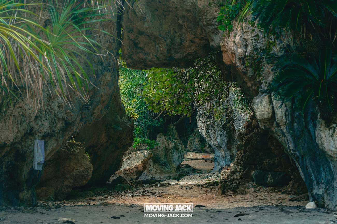 Grote rotsachtige boog, omgeven door weelderige groene planten, waar zonlicht doorheen filtert. Zandgrond en rotsen zijn zichtbaar, samen met een handdoek die links hangt – een herinnering aan de beste stranden die Okinawa te bieden heeft. "Moving Jack" watermerk middenonder. -copyright-moving-jack.com