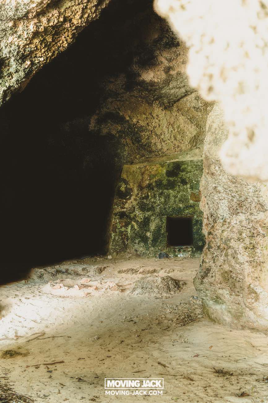 Een grot met ruwe stenen muren en een kleine rechthoekige opening in de achterwand, gedeeltelijk verlicht door zonlicht dat van buitenaf naar binnen stroomt – net als de verborgen plekjes bij de beste stranden van Okinawa. De grond is bedekt met aarde en stenen. -copyright-moving-jack.com