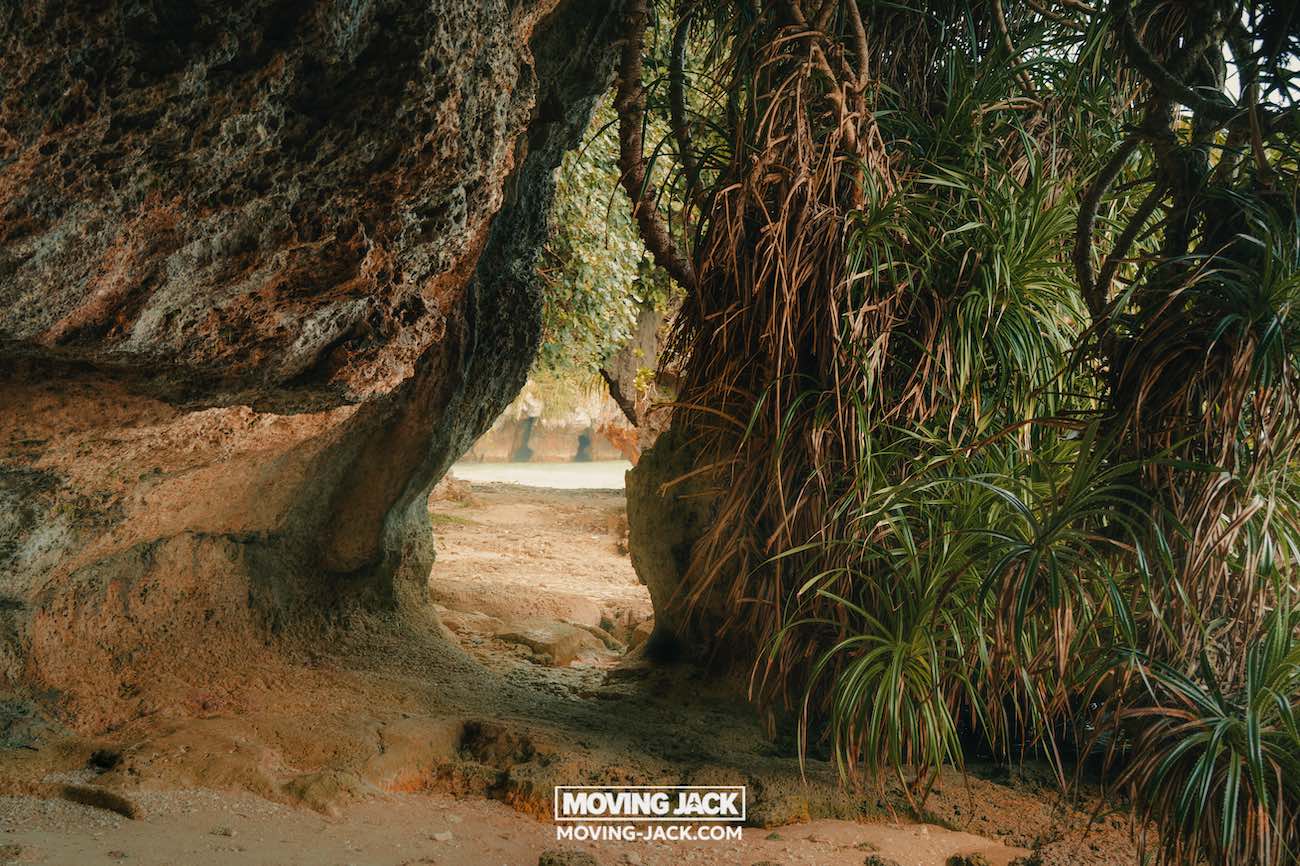 Een zandpad slingert tussen grote rotsachtige kliffen en weelderige groene planten en leidt je naar een zonnig strand met sprankelend water, waar de natuurlijke, tropische schoonheid van de beste stranden van Okinawa wordt vastgelegd. Onderaan staat het logo van "moving jack". -copyright-moving-jack.com