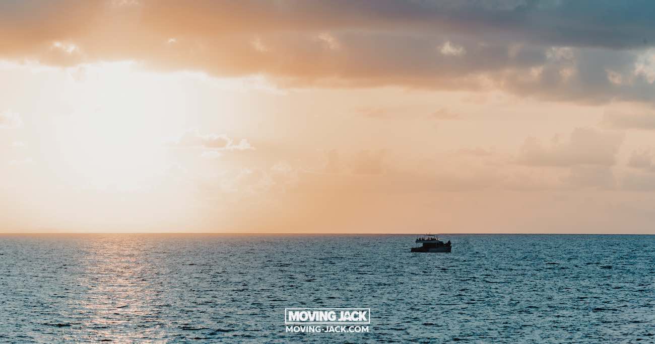 Een boot drijft op kalm oceaanwater onder een bewolkte zonsonderganghemel nabij de beste stranden van Okinawa, met de zon die een warme gloed en glinsterende weerspiegelingen over de zee werpt. -copyright-moving-jack.com