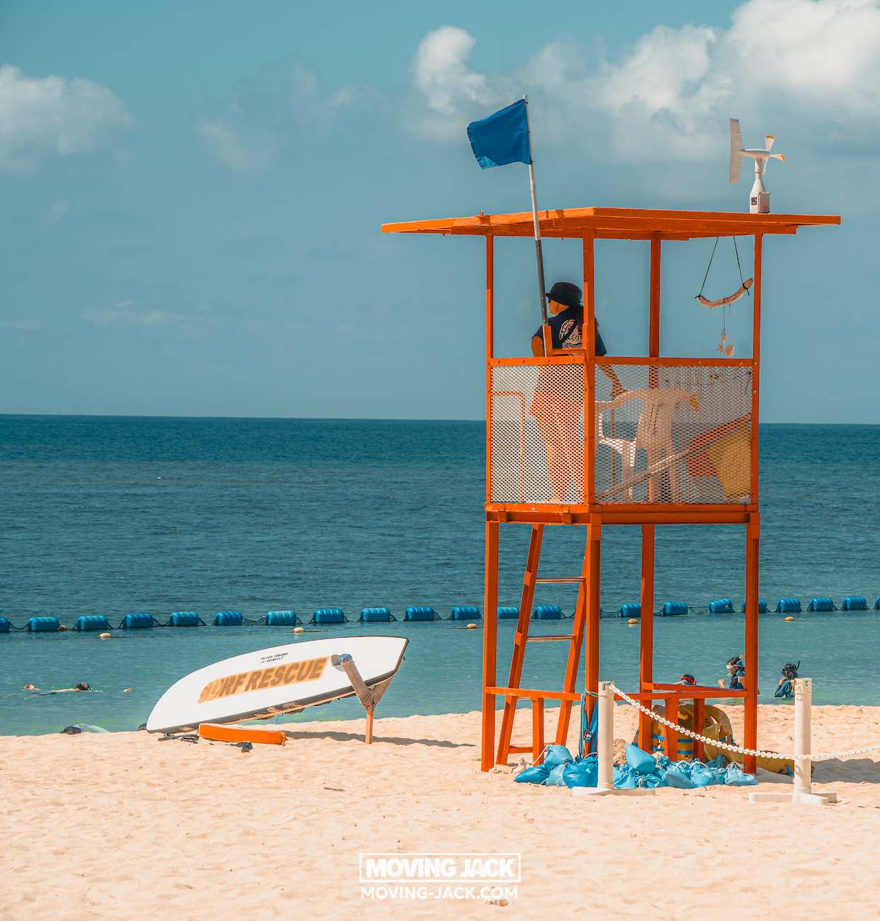 Een strandwachter staat op een oranje toren te waken over een zandstrand en een blauwe oceaan – een van de mooiste stranden die Okinawa te bieden heeft. Zwemmers genieten van het water, terwijl een wit reddingsbord met het opschrift "infrescue" vlakbij rust onder een gedeeltelijk bewolkte hemel. -copyright-moving-jack.com
