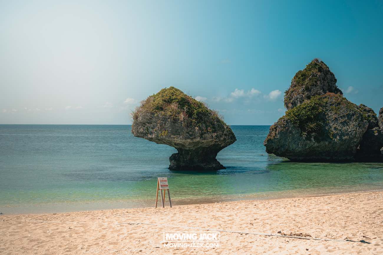 Een zandstrand met helder turquoise water, unieke rotsformaties bedekt met groen in de ondiepe zee en een klein waarschuwingsbordje vlak bij de kust onder een stralend blauwe lucht: een van de beste stranden die Okinawa te bieden heeft. -copyright-moving-jack.com