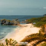 Een rustig zandstrand met turquoise water, rotsformaties en weelderig groen – een van de beste stranden die Okinawa te bieden heeft. Een paar mensen slenteren langs de kust en een kleine blauwe luifel hangt vlak bij het water onder een heldere, zonnige hemel. -copyright-moving-jack.com