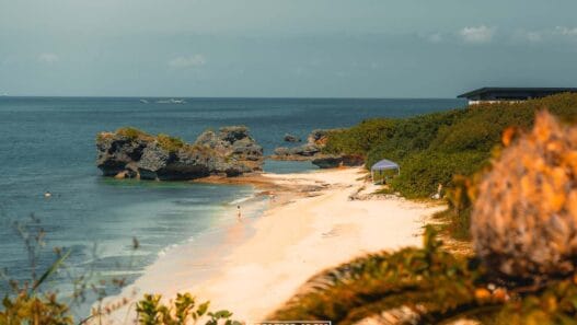 Een rustig zandstrand met turquoise water, rotsformaties en weelderig groen – een van de beste stranden die Okinawa te bieden heeft. Een paar mensen slenteren langs de kust en een kleine blauwe luifel hangt vlak bij het water onder een heldere, zonnige hemel. -copyright-moving-jack.com