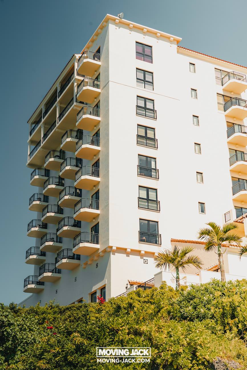 Een hoog, wit appartementengebouw met meerdere balkons en zwarte balustrades staat tegen een helderblauwe lucht, omgeven door groene struiken en palmbomen – perfect voor wie op zoek is naar gemakkelijke toegang tot de beste stranden die Okinawa te bieden heeft. -copyright-moving-jack.com