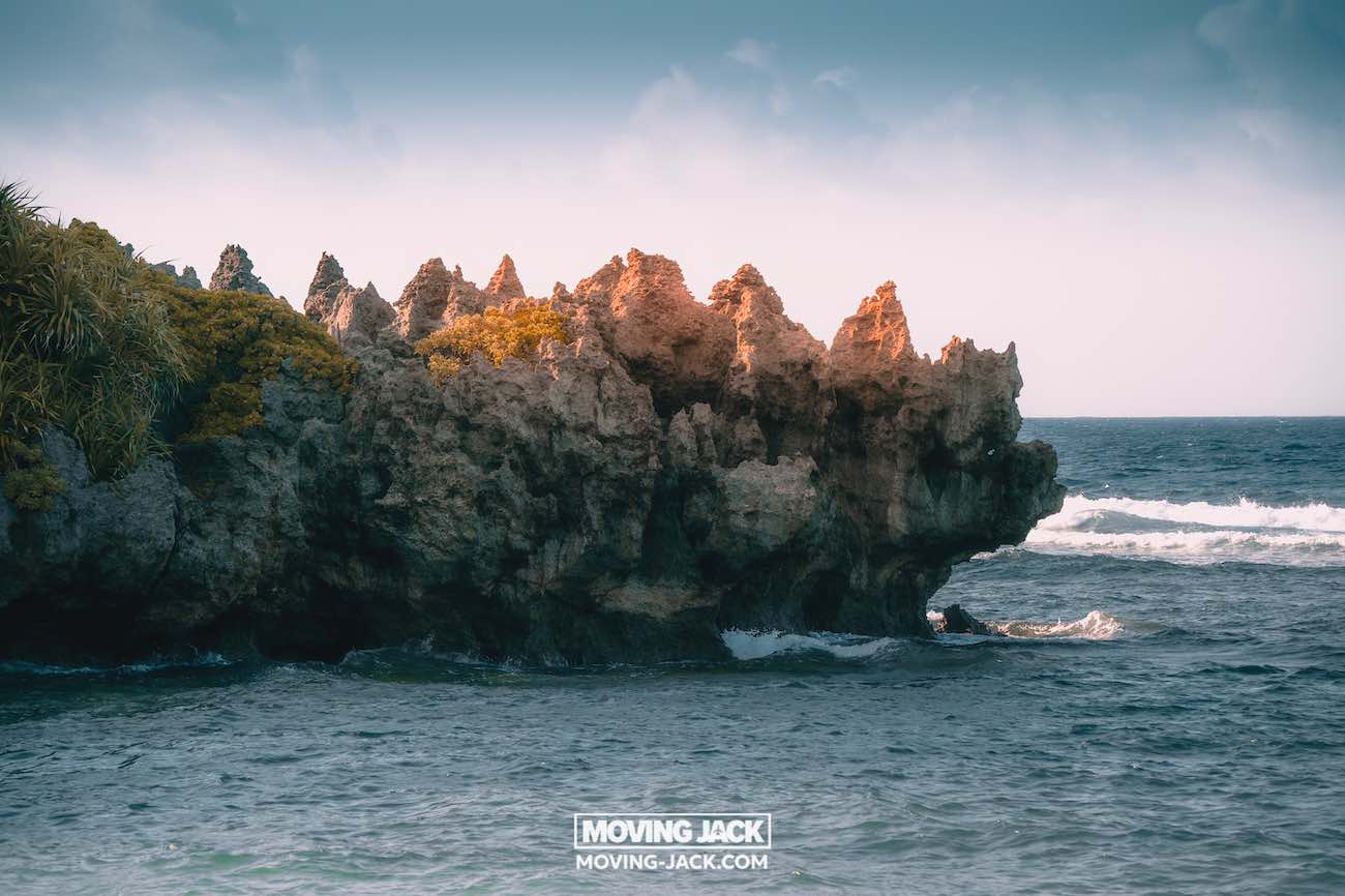 Een grillige rotsformatie met weinig groen steekt uit in de blauwe oceaan, golven beuken onder een gedeeltelijk bewolkte hemel. Een watermerk vermeldt "moving jack moving-jack.com". Ontdek dit soort landschappen in de buurt van enkele van de beste stranden die Okinawa te bieden heeft. -copyright-moving-jack.com
