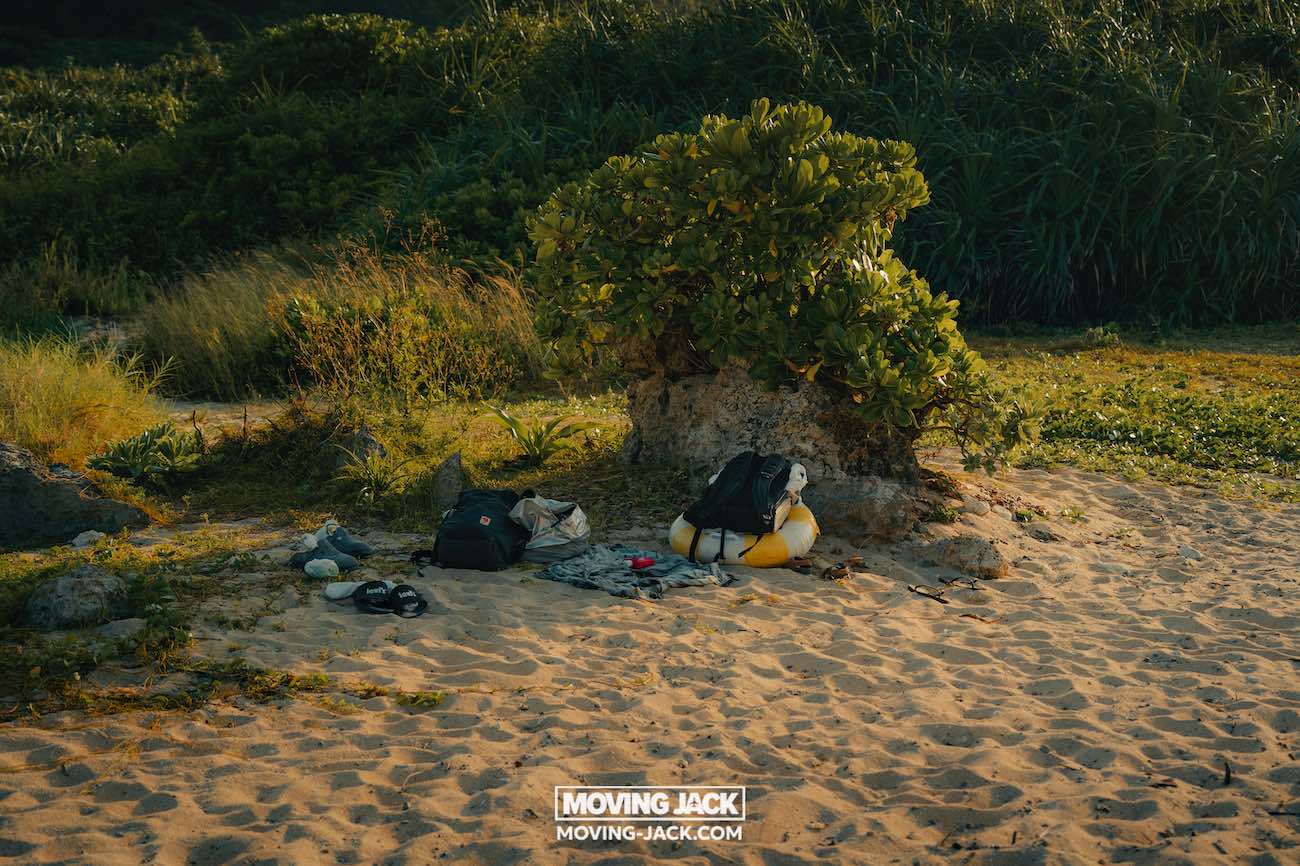 Rugzakken, kleding en uitrusting liggen op zanderige grond naast een groene struik en rots, met weelderige vegetatie op de achtergrond in warm zonlicht. Ze leggen een avontuur vast nabij de beste stranden die Okinawa te bieden heeft. Het logo van "moving jack" verschijnt onderaan. -copyright-moving-jack.com