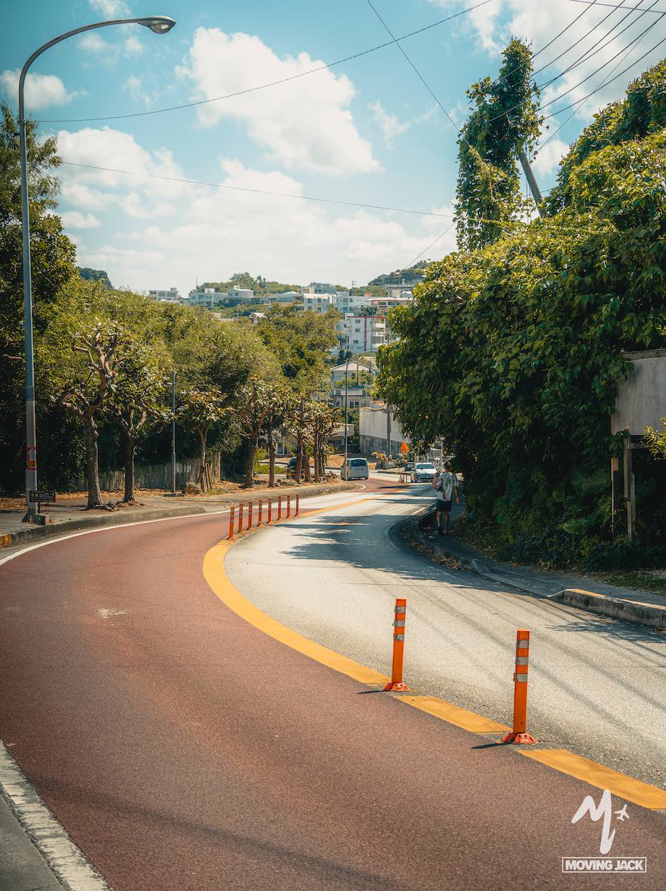 Een bochtige weg met bomen en groen loopt door een zonnig stedelijk gebied en leidt naar Senaha Beach, een van de mooiste stranden van Okinawa.