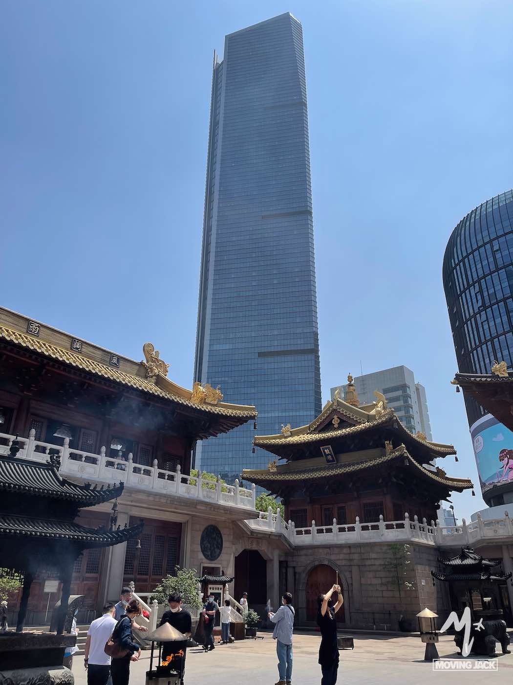 Mensen verzamelen zich op de binnenplaats van een traditionele Chinese tempel, met sierlijke daken op de voorgrond en een moderne glazen wolkenkrabber die hoog oprijst tegen de helderblauwe hemel daarachter. -Copyright-moving-jack.com