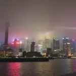 A nighttime cityscape of shanghai’s skyline with brightly lit skyscrapers and the oriental pearl tower, shrouded in fog, reflecting on the river. People are holding umbrellas in the lower right corner. -copyright-moving-jack. Com