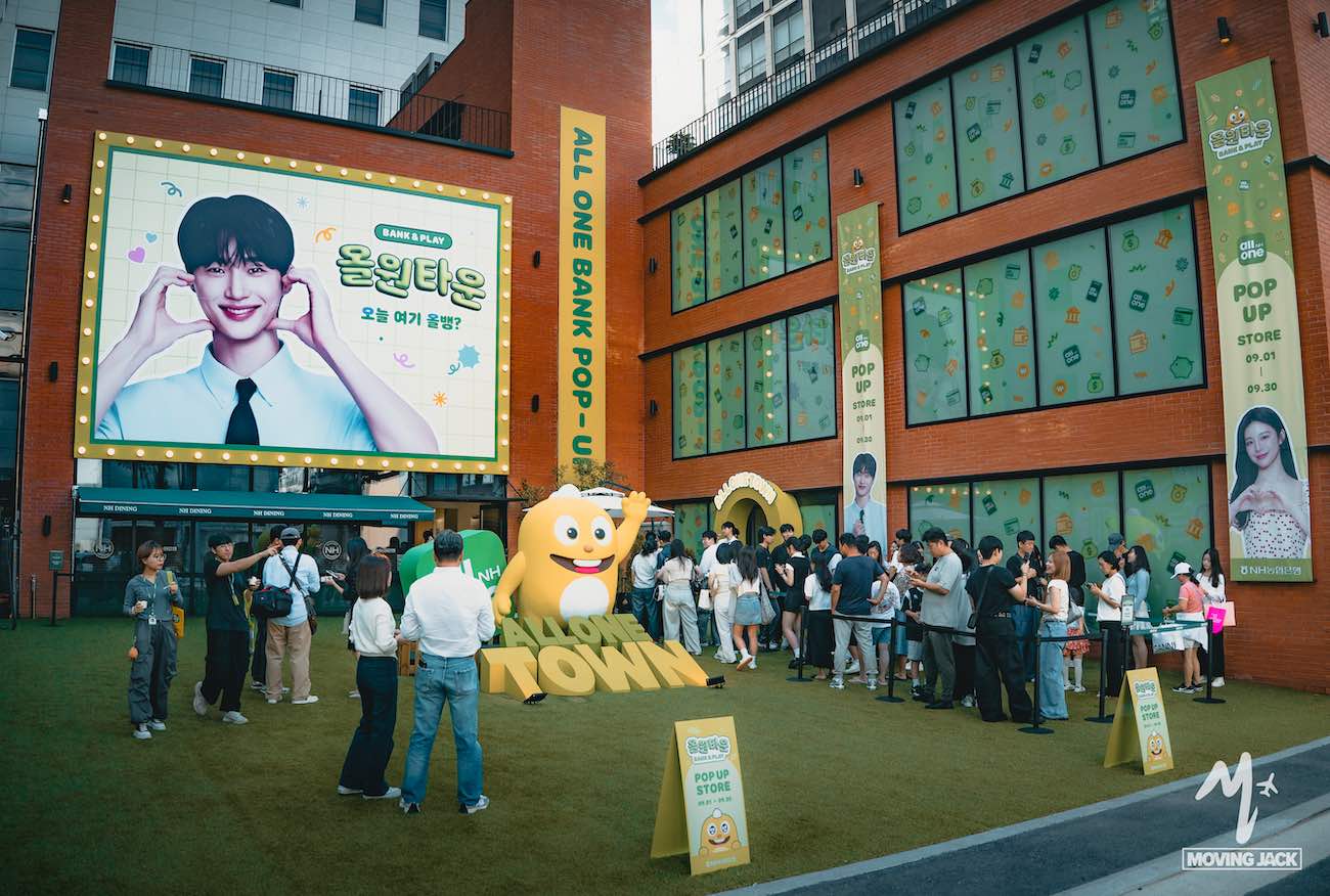 Mensen staan in de rij voor een kleurrijke pop-upwinkel met cartoonmascottes en grote posters met lachende figuren. Het gebouw is versierd met groene en gele decoraties en borden met de teksten "all one bank pop-up" en "holown town". -copyright-moving-jack.com