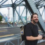 A smiling man with a backpack and camera stands on a steel bridge, with modern skyscrapers and the oriental pearl tower visible in the background on a cloudy day. -copyright-moving-jack. Com