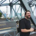 A smiling man with a backpack and camera stands on a steel bridge, with modern skyscrapers and the oriental pearl tower visible in the background on a cloudy day. -copyright-moving-jack. Com