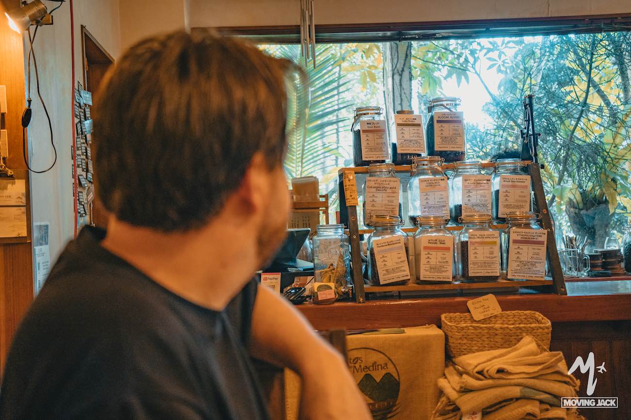 A man with dark hair sits indoors, facing a display of labeled glass jars arranged on shelves in front of a window with greenery outside—perhaps planning his okinawa itinerary as towels and bags sit stacked on the wooden counter nearby. -copyright-moving-jack. Com