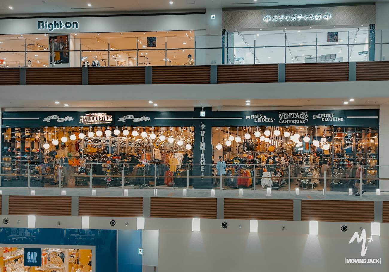 A vintage clothing store with warm hanging lights and a decorative interior sits on the second floor of a shopping mall—perfect for adding a unique stop to your okinawa itinerary, with other stores and shoppers visible nearby. -copyright-moving-jack. Com