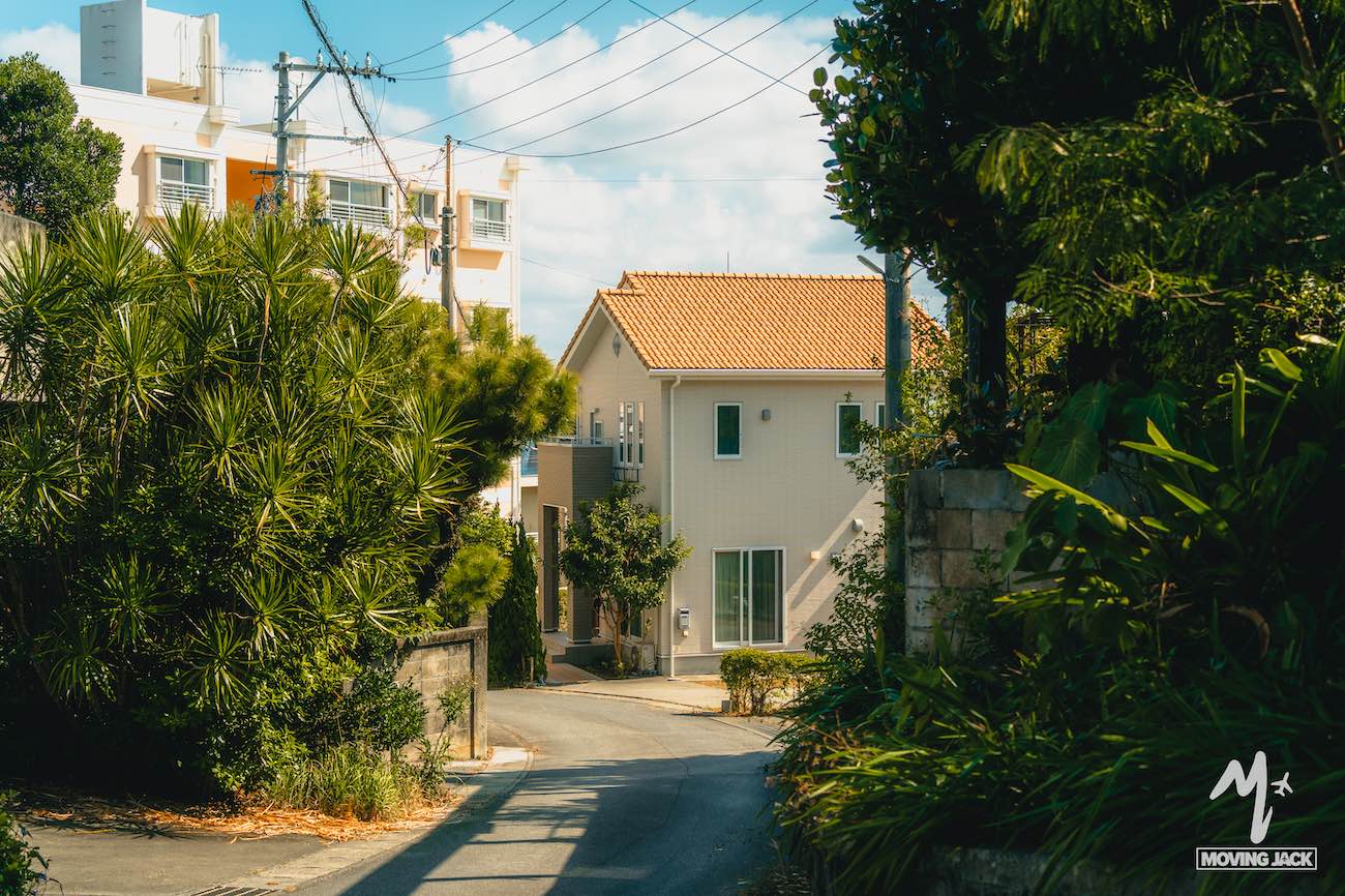 A quiet residential street with lush green plants and trees on both sides, leading to a beige house with a tiled roof—an inviting scene for any okinawa itinerary, with power lines and an apartment building under a partly cloudy sky. -copyright-moving-jack. Com