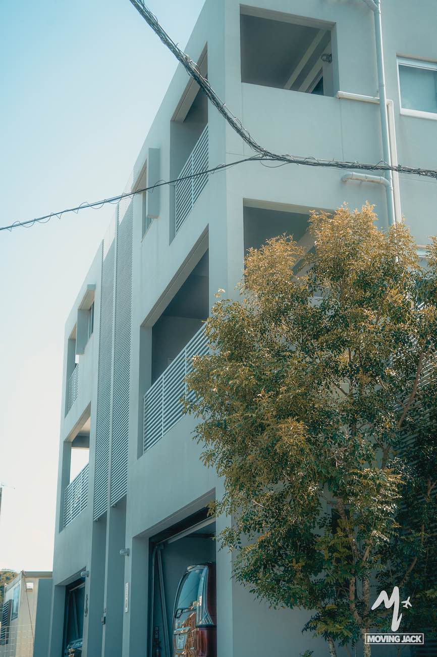 A modern, three-story gray building with open balconies and garage spaces on the ground floor. A tree with green leaves stands in front under a clear sky—perfect inspiration for your okinawa itinerary. The moving jack logo appears at the bottom right. -copyright-moving-jack. Com