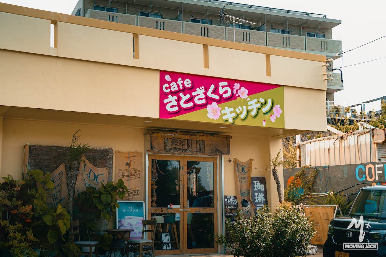 A yellow cafe with a wooden door and plants outside, perfect for your okinawa itinerary. A pink and green sign above the entrance reads “cafe さとざくら キッチン. ” residential buildings are visible in the background. -copyright-moving-jack. Com